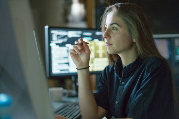 Woman monitors dark office A young woman is seated at a desk surrounded by monitors displaying data, she is contemplating in this dark, moody office. journalist stock pictures, royalty-free photos & images