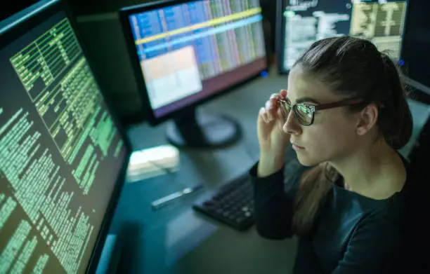 A young woman is seated at a desk surrounded by monitors displaying data, she is contemplating in this dark, moody office.