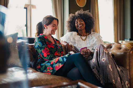 Two beautiful women are sitting comfortably in a luxury hotel, laughing. One of the women is holding a mobile phone in her hand.