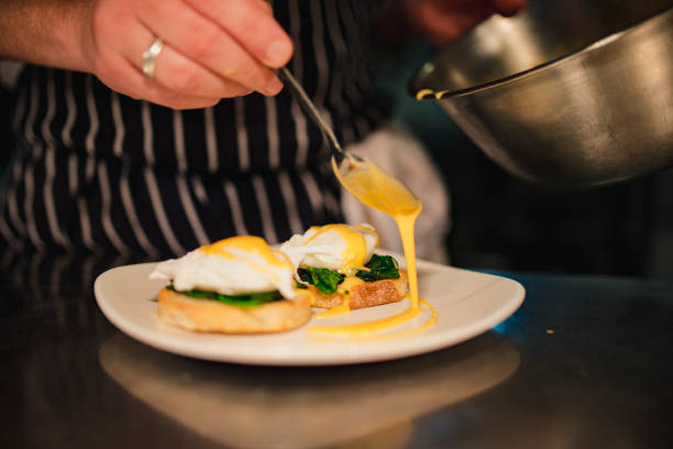 Breakfast Perfection A surface-level shot of poached eggs on toast on a plate, being covered in hollandaise sauce by an unrecognisable hotel chef. hollandaise sauce stock pictures, royalty-free photos & images