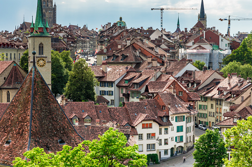 Color image depicting the historic old town of Bern, the capital city of Switzerland. The town is full of church spires, old tiled roofs and cobblestone streets. Lush green trees abound, and the image is offset by a blue sky and cloudscape. Room for copy space.