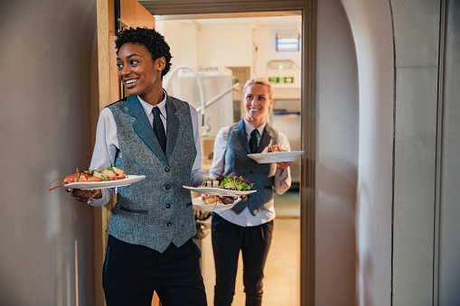 Two waiters are coming out of a hotel kitchen while holding two dishes each, ready to serve to guests.