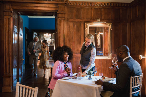 What a Lovely Meal! A group of well-dressed friends dining in a luxury hotel, being served a meal by a waitress. Other guests are walking towards the restaurant in the background, ready to dine. exclusive dinner stock pictures, royalty-free photos & images