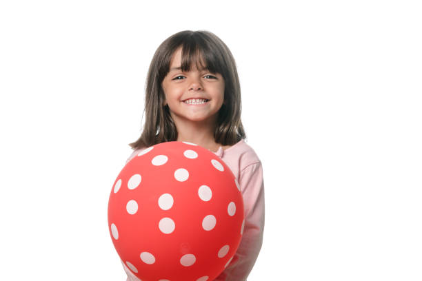 Portrait Of Cute Girl Holding Balloons While Standing On Grassy Field Against Fence