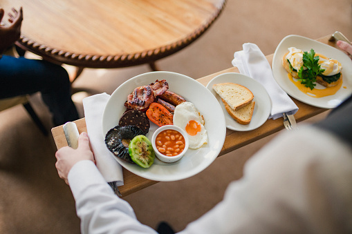 A waiter is serving a breakfast tray with a full English breakfast including a fried egg, bacon, sausage, mushrooms and beans on a white plate and poached eggs/