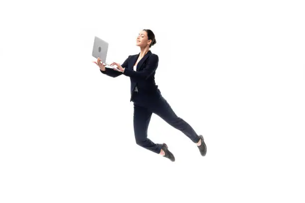 young cheerful businesswoman using laptop while levitating isolated on white