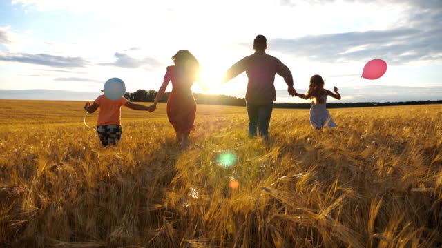 Young couple of parents with two children holding hands of each other and running through wheat field at sunset. Happy family jogging among barley meadow and enjoying nature together. Slow motion