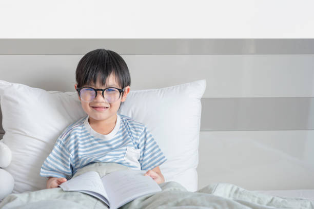 happy asian boy happy child little asian boy smile with holding glasses reading a books on the bedroom in room at home. family activity concept. korean doll stock pictures, royalty-free photos & images