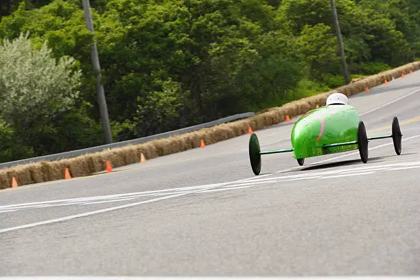 Aerodynamic green master-class soapbox derby car and driver stopped in action coming down the track during competition.  In the master class the driver is in a prone position in the cockpit peering above cowling.  Soap box derby car crosses the finish line in abstract motion blur.  The Soap Box Derby is a youth car racing program which has been run in the United States since 1934. World Championship finals are held each July at Derby Downs in Akron, Ohio. Race cars are unpowered, relying entirely upon gravity to move downhill.  Early cars were constructed of wood from crates and other scrap sources, with wheels from baby buggies and wagons.  Today, using standardized wheels with precision ball bearings, modern racers start on a ramp at the top of a hill, and attain speeds of up to 35 miles per hour.  Irondequoit, New York, 2008.