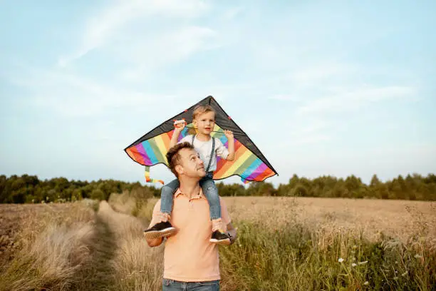 Portrait of a happy father and young son on the shoulders with colorful air kite on the blue sky background. Concept of a happy family and summer activity