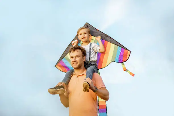 Portrait of a happy father and young son on the shoulders with colorful air kite on the blue sky background. Concept of a happy family and summer activity