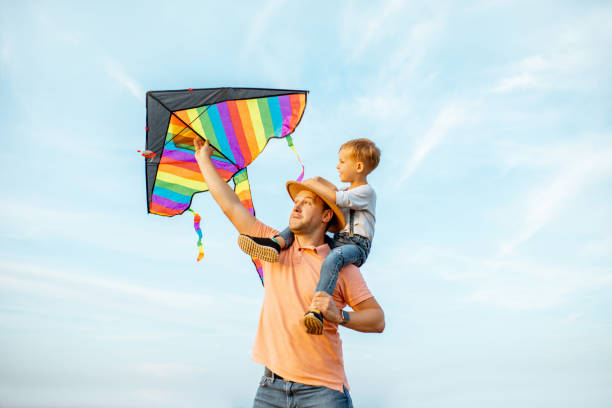 Father and son with air kite outdoors Portrait of a happy father and young son on the shoulders with colorful air kite on the blue sky background. Concept of a happy family and summer activity kite sailing stock pictures, royalty-free photos & images