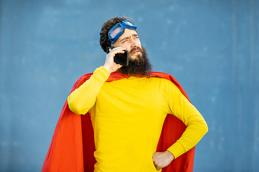 Portrait of a man like a superhero in colorful clothes and pilot's glasses holding smart phone while standing on the blue background