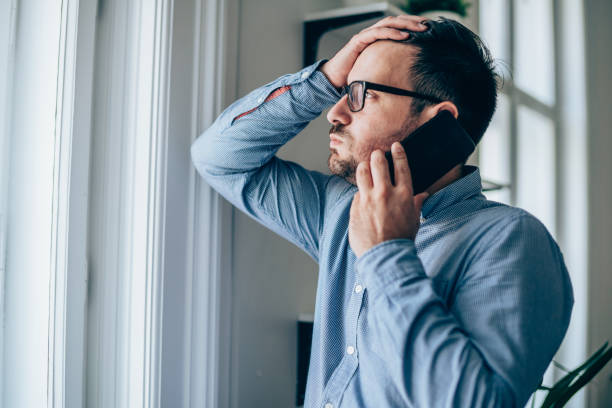 Frustrated businessman using phone Worried businessman with hand on forehead talking on mobile phone in his office. edgy man stock pictures, royalty-free photos & images