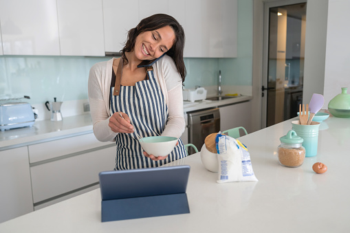 Woman multi tasking at home cooking, looking at a recipe on tablet and on a phonecall smiling very happy