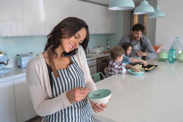 istock Young mother multi tasking using smartphone and mixing something in bowl while father and kids cook at background 1169630637