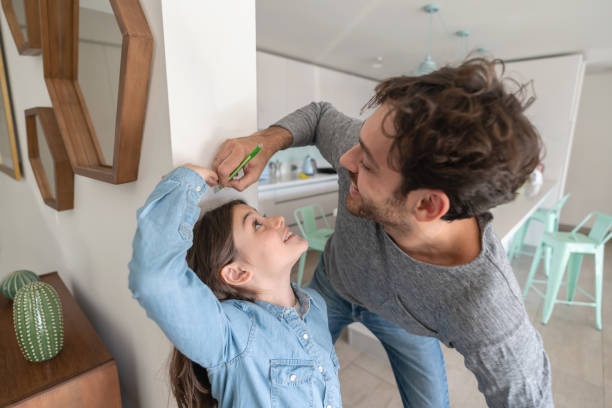 Happy father measuring daughter against the walll while she cheats with her height both smiling