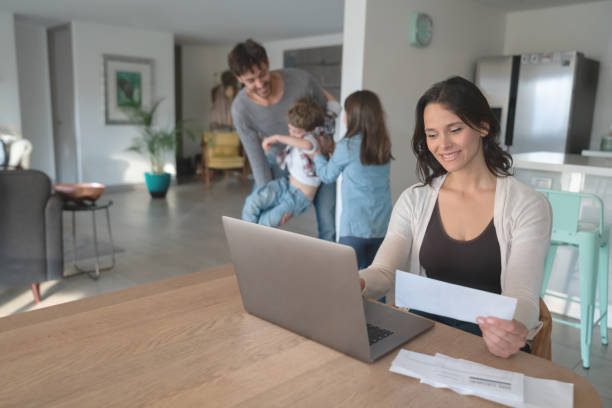 istock Beautiful young mother paying bills online using a laptop and father playing with his two kids at background 1169630708