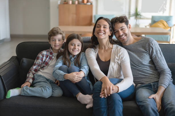 cute latin american coupe with two children at home sitting on couch smiling at camera and mom with a toothy smile - offspring child toothy smile beautiful imagens e fotografias de stock