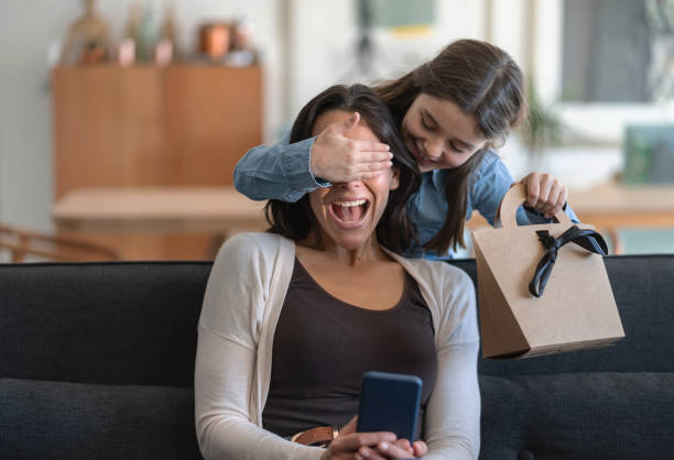 excited mom being surprised by daughter who is covering her eyes and holding a gift for mother's day - mother gift imagens e fotografias de stock