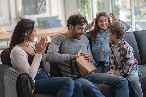 Excited children and mother waiting for daddy to open a present for father's day while sitting on couch at home - Celebration concepts