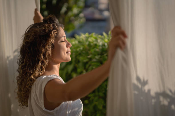 donna tranquilla che guarda attraverso la finestra della sua camera da letto al mattino - waking up window women morning foto e immagini stock