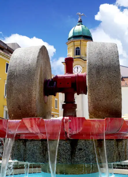 Millstone in a fountain and a city clock on a cultural monument in the background. Cultural and traditional heritage of the Croatian city of Rijeka
