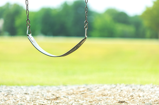 public park with empty swing on playground of public grassy outlined with trees blurred in background (Shot with Canon 5DS 50.6mp photos professionally retouched - Lightroom / Photoshop - original size 5792 x 8688 downsampled as needed for clarity and select focus used for dramatic effect)