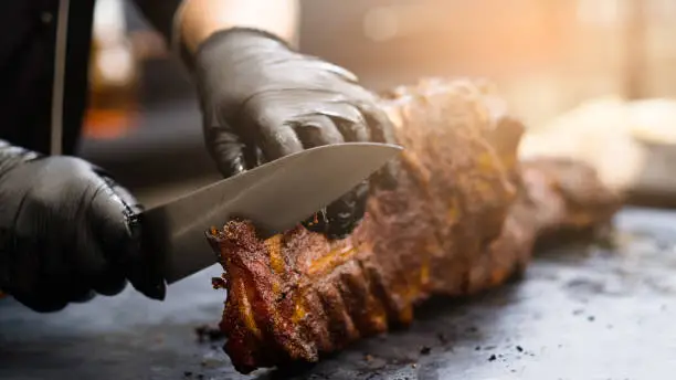 Grill restaurant kitchen. Cropped shot of chef in black cooking gloves using knife to cut smoked pork ribs.