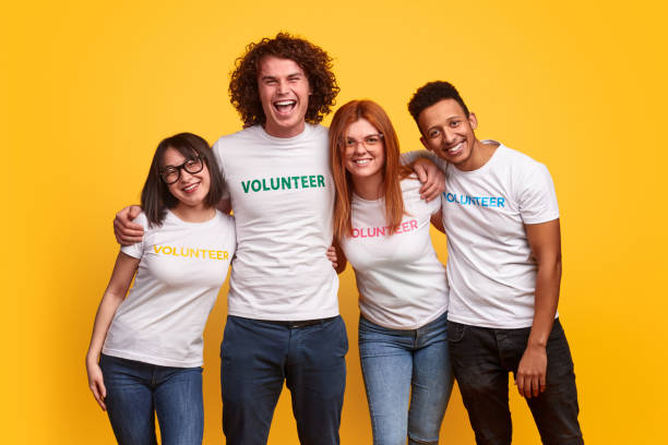 Happy multiethnic volunteers hugging each other Group of cheerful multiracial young people in volunteer T-shirts smiling and embracing each other against yellow background altruism photos stock pictures, royalty-free photos & images