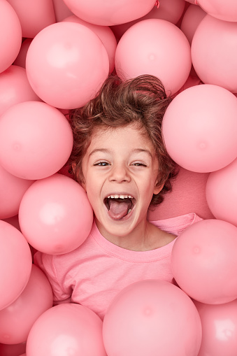 From above happy little boy showing tongue and looking at camera while playing in heap of pink balloons