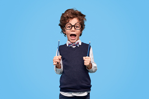 Excited little nerd in round glasses and school uniform screaming and holding two pencils while being ready go back to school against blue background