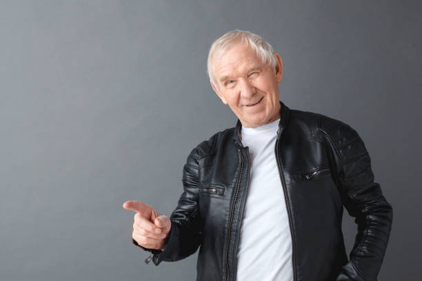 istock Senior man in leather jacket standing isolated on gray pointing camera winking playful 1169656586