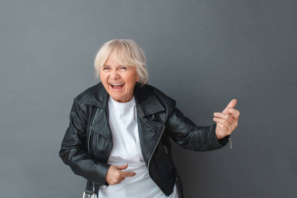 istock Senior woman in leather jacket studio standing isolated on gray playing imaginary guitar laughing playful close-up 1169656591
