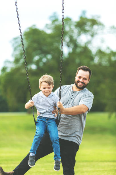 père millénaire jouant avec le fils balançant sur le balançoire à un terrain de jeu de stationnement public ayant l'amusement ensemble - summer recreation photos et images de collection