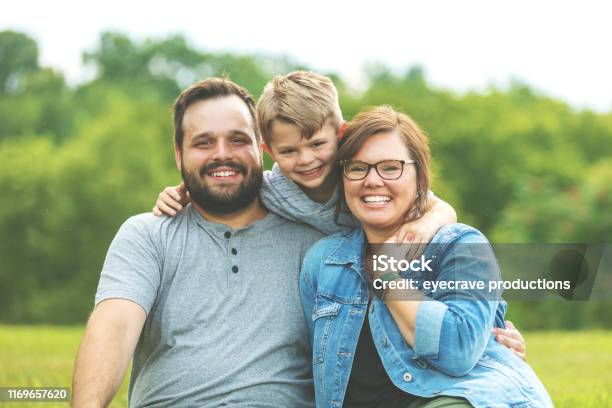 Millennial Father And Mother With Autistic Child In Public Lush Green Park Sitting On The Grass Smiling Pose For Family Portrait Stock Photo - Download Image Now