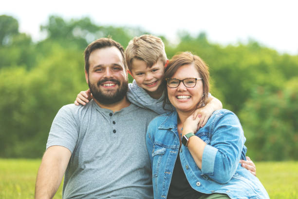 millennial father and mother with autistic child in public lush green park sitting on the grass smiling pose for family portrait - child offspring women posing imagens e fotografias de stock