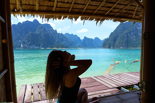 Asian woman sitting and relaxing on bamboo raft with lake and limestone mountain range background in Surat Thani, Thailand.