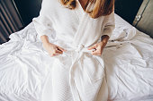 Cropped shot view of young woman sitting on the bed and tying up belt of white bathrobe before taking a bath or spa massage.