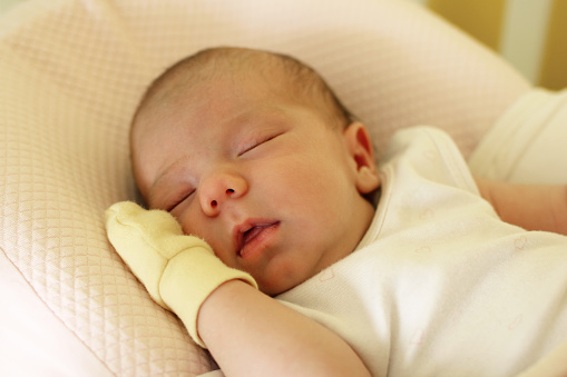 Cute newborn baby with open mouth and yellow mitten on a hand is sleeping on her bed.