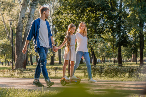 Side view of young smiling parents walking in park with their cute little girl riding hoveboard Public Park, Segway, Summer, Ukraine, Family hoverboard stock pictures, royalty-free photos & images
