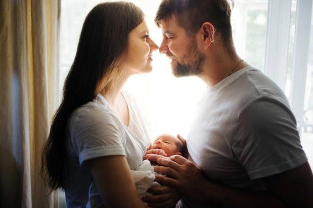 Father and mother with a newborn son against the background of the window in the backlight Father and mother with a newborn son against the background of the window in the backlight. New young multi-generation family. Breast-feeding cute pregnant woman with gift stock pictures, royalty-free photos & images