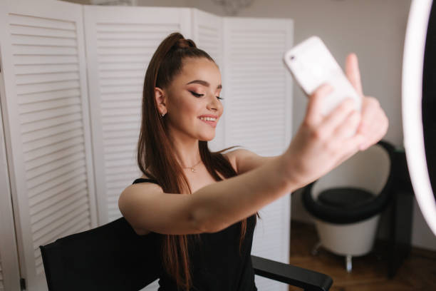 istock Young attractive model make a selfie after makeup in beauty studio. She use a ring lapm for photo. Background of white folding screen 1169724554