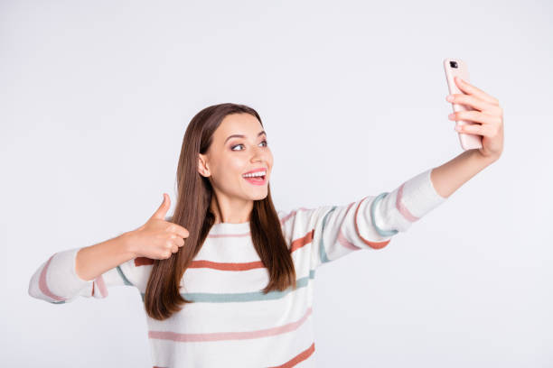 istock Pretty lady making selfie on new telephone raising thumb up wear striped pullover isolated white background 1169724882