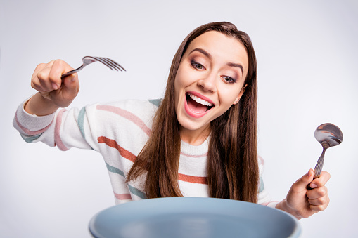 Hungry lady holding fork and spoon can't wait to start, eating wear striped pullover isolated white background