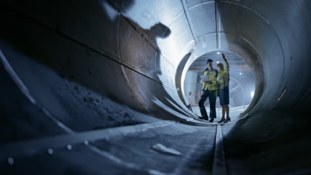 Two Heavy Industry Engineers Walking Inside Pipe, Use Laptop, Have Discussion, Checking Design. Construction of the Oil, Natural Gas and Biofuels Transport Pipeline. Industrial Manufacturing Factory