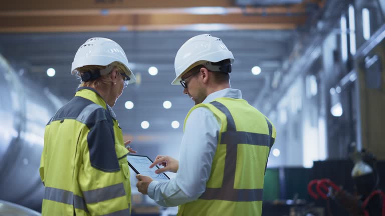 Two Heavy Industry Engineers Stand in Pipe Manufacturing Factory, Use Digital Tablet Computer, Have Discussion. Construction of Oil, Gas and Fuels Transport Pipeline. Back View Sparks Flying