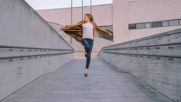 istock Cheerful and Happy Young Woman Actively Dancing and Jumping While Walking Down a Concrete Urban Path. She's Wearing a Long Brown Coat. Scene Shot Next to Business Center. Day is Bright. 1169740980