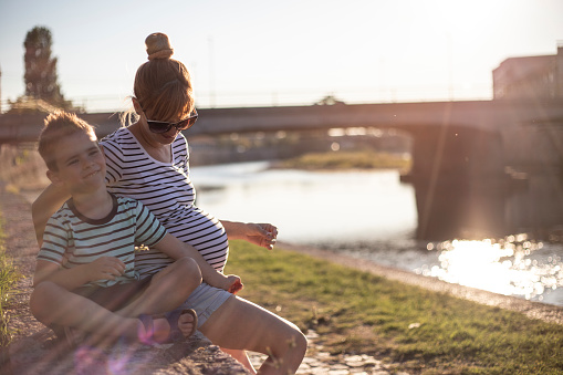 Young pregnant woman sitting by the river and enjoying with her five years old son.