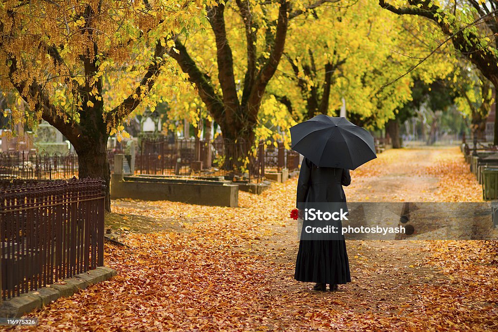 Mourning Woman wearing black in cemetery in fall A lone woman in mourning in a cemetery in fall (autumn), dressed in black and carrying a black umbrella and a red flower. The central path of the cemetery is covered with orange leaves and the surrounding trees glow yellow and orange and enhance the emotion of the image. Widow Stock Photo